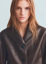 Chaqueta corta de piel sintética en color marrón, de diseño recto con cuello en V, solapas de muesca, manga larga, dos bolsillos laterales y cierre frontal de botones.