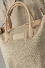 Cartera tipo bolso de mano confeccionada en lana fina sin teñir, con detalles en cuero marrón oscuro en las asas y correa ajustable. Incluye cierre superior y etiqueta de cuero con logo.