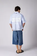 Camisa de manga corta a rayas beige y blancas, de corte boxy y liviana, con cuello camisero, cierre frontal de botones y pequeño texto bordado "BUILD IT BOLD" en el pecho.