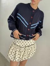 Saco de punto color crema con detalles en crochet verde, cierre frontal con botones y mangas largas.