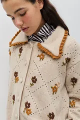 Saco de punto color beige con bordados artesanales de flores en tonos marrones y ocres. Presenta cuello con detalle de volados en color ocre, cierre frontal con botones forrados y silueta amplia.