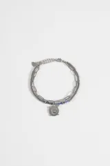 Pulsera de acero quirúrgico plateado de doble tira, una de cadena eslabonada y otra con cuentas de metal y piedras azules. Cuenta con un dije colgante circular con diseño de ojo y detalles grabados.