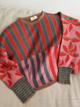 Sweater de tejido de punto con silueta oversize, cuello redondo y mangas largas abullonadas. Presenta un diseño patchwork con rayas verticales en tonos rojo, rosa y gris verdoso en el cuerpo, y un estampado floral en tonos rosa y rojo con detalles dorados en las mangas. Los puños y el cuello tienen un ribete a rayas horizontales.