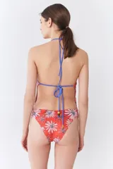 Traje de baño entero con escote en V y tirantes finos, estampado con diseño de flores tipo margarita sobre fondo rojo. Presenta un sistema de tiras cruzadas en la espalda y terminaciones artesanales en resina.