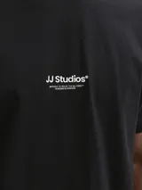 Remera negra de manga corta con cuello redondo y calce relajado, confeccionada en jersey de algodón orgánico. Presenta un pequeño estampado tipográfico en el centro del pecho con el texto 'JJ Studios'.
