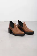 Botín asimétrico de estilo Chelsea color bordó, confeccionado en cuero vacuno, con paneles elásticos laterales negros, puntera cuadrada, suela de plataforma y tacón de bloque cuadrado.
