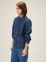 Camisa de denim liviano color azul, con cuello mao y detalles de volados fruncidos en el cuello y la botonera frontal. Presenta mangas largas con puños rectos y corte relajado.