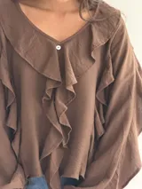 Blusa blanca de corte holgado con escote en V, decorada con volados frontales y en las mangas. Presenta mangas largas con puños elásticos y cierre frontal con botones.