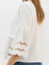 Camisa negra de poliéster con cuello camisero, escote en V y cierre frontal de botones. Cuenta con mangas anchas de corte acampanado y detalles de bordado suizo calado en las mangas y en el ruedo inferior.