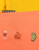 Neceser rectangular de tela color ladrillo con cierre superior y tres bordados pequeños: una hoja de arce, una taza de té y un croissant.