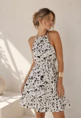 Vestido corto sin mangas con cuello halter, falda con volados y cinturón ajustable en la misma tela. Presenta un diseño estampado en tonos blanco y negro.