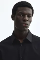 Camisa de vestir negra con cuello inglés, tapeta clásica y mangas largas con puños ajustables. Presenta un corte entallado que se ajusta al cuerpo y bajo levemente redondeado.