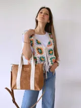Cartera tipo tote de cuero color camel con detalles de bandas verticales en color crema. Cuenta con doble asa de mano, correa larga ajustable y extraíble, y cierre superior. Incluye etiqueta de cuero con logo grabado en el frente.