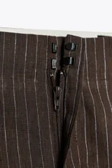 Pantalón de tiro medio confeccionado en hilatura con mezcla de lino a rayas verticales marrones y blancas. Presenta bolsillos laterales ocultos en costura, detalle de pliegues en el bajo y cierre frontal con cremallera, botón y ganchos metálicos.