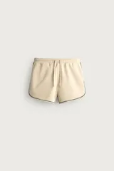Bermuda de corte relajado confeccionada en tejido de algodón, color beige con vivos en contraste color verde oscuro. Cuenta con cintura elástica ajustable con cordón, bolsillos frontales y bolsillos traseros de plastrón.