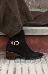 Bota corta de cuero negro con taco cuadrado de madera y punta fina. Presenta una tira decorativa con hebilla metálica dorada en el tobillo.