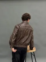 Campera estilo bomber de cuero sintético en color marrón con efecto desgastado. Presenta cuello camisero, cierre frontal con botones metálicos, bolsillos frontales con solapa y cintura elástica.