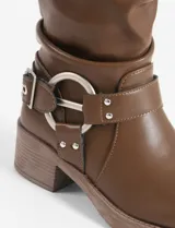 Bota corta de cuero vegano color marrón, con diseño de caña arrugada, hebilla decorativa en la parte superior y arnés metálico con hebilla en el tobillo. Cuenta con un taco cuadrado de altura media.