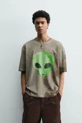 Camiseta de corte relajado con cuello redondo y manga corta. Presenta un efecto de lavado desgastado en tonos marrones y un estampado frontal de una cabeza de alienígena en color verde vibrante.