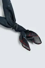 Pañuelo cuadrado de algodón color verde oliva con un patrón de pequeñas cruces blancas distribuidas por toda la superficie. El borde presenta un ribete con un patrón de cuadros en tonos oscuros (negro, azul marino) y líneas finas blancas.