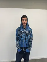 Buzo tipo hoodie tejido de punto grueso con cierre frontal completo hasta la capucha. Presenta un diseño gráfico abstracto en tonos azules y negros, con tipografía estilo gótico en el frente y las mangas. Cuenta con puños y ruedo acanalados en color negro, y etiqueta de marca en la manga.