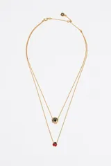 Collar doble de cadena fina dorada con cierre de mosquetón. La cadena superior cuenta con un dije circular dorado con un corazón negro central y grabado de logo, mientras que la cadena inferior presenta un dije en forma de mariquita roja esmaltada con detalles negros.