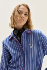 Camisa de corte holgado con diseño de rayas verticales en azul y blanco. Presenta cuello clásico y cartera frontal con detalles bordados en hilo rojo. Incluye un pequeño bordado de estrellas en el pecho y mangas largas con puños estructurados.