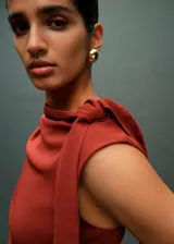 Vestido midi de fiesta color terracota, con cuello barco y diseño sin mangas. Presenta detalles fruncidos en la cintura y un detalle de nudo en el hombro, con un corte entallado y cierre posterior.