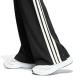 Pantalón de mujer Adidas tipo jogging en color negro, con diseño de malla transpirable, corte ancho y franjas blancas laterales con las clásicas tres tiras de la marca. Cuenta con cintura elástica con cordón de ajuste y logo bordado en la pierna.