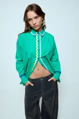 Camisa de algodón color verde, de corte oversize y manga larga. Presenta detalles de cintas onduladas en contraste sobre la botonadura frontal y en los puños, además de un ribete en el cuello.
