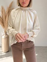 Blusa de tela fluida color beige, con cuello alto fruncido y mangas largas amplias terminadas en puños elastizados con detalle de fruncido.