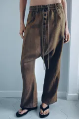 Pantalón tipo baggy de tiro bajo, confeccionado en mezcla de lino. Presenta cintura elástica con cordón ajustable, cierre frontal con botones y falsos bolsillos de vivo en los laterales.