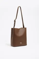 Bolso de hombro Bimba y Lola modelo Carlino, de estructura rígida y color marrón, confeccionado en cuero. Cuenta con asa regulable y logo de la marca estampado en plateado en el frente.
