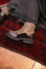 Bota corta de estilo urbano con diseño bicolor, combinando paneles de gamuza en color arena y cuero negro en la puntera y el talón. Cuenta con suela track de goma negra y cierre lateral.