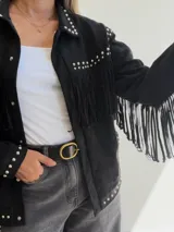 Chaqueta de efecto gamuza color negro, con diseño de flecos en el pecho y mangas. Presenta detalles de tachas metálicas plateadas en el cuello, hombros, puños y ruedo. Cuenta con cierre frontal de botones y bolsillos funcionales.