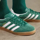 Championes Adidas Gazelle Indoor de gamuza color verde, con las icónicas tres tiras laterales en blanco y suela de caucho color caramelo. Presenta el nombre del modelo en letras doradas y detalle en el talón en color blanco.