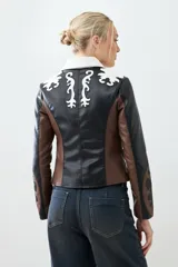 Chaqueta corta de cuero sintético color negro, marrón y blanco, con cuello de piel sintética blanca, cierre frontal con cremallera y bordado en las mangas.
