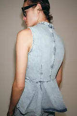 Chaleco de denim claro con efecto lavado, cuello mao, sin mangas y corte peplum con pliegues en la parte inferior. Cierre frontal con botones de alamares negros.