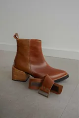 Bota corta de cuero negro con acabado combinado mate y charol. Presenta una tira decorativa con tachas metálicas y hebilla en el tobillo, tacón cuadrado robusto y suela con plataforma.