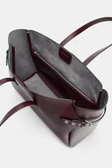 Bolso tipo shopper de gran tamaño en color bordó, con diseño estructurado y detalles de correas laterales con ojales metálicos y hebillas. Cuenta con doble asa de mano y cierre superior mediante imán.