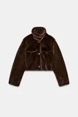 Abrigo corto tipo bomber con textura de pelo sintético, cuello subido, manga larga y cierre frontal con botones. Presenta bolsillos decorativos con solapa en el frente.