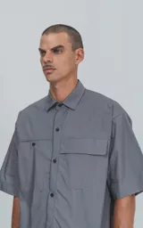Camisa de manga corta en color gris, confeccionada en popelina de algodón. Presenta un corte oversize, cuello clásico, cierre frontal con botones y dos bolsillos delanteros, uno de ellos con solapa.