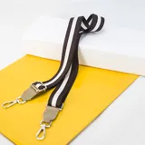 Correa de tela negra con franjas color beige, terminaciones en cuero sintético negro y mosquetones metálicos plateados.