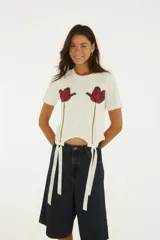 Remera blanca de manga corta y cuello redondo, confeccionada en algodón, con estampado frontal de dos tulipanes rojos y detalles de lazos de satén en el dobladillo.