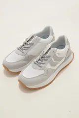 Zapatilla deportiva de diseño urbano, combinando paneles de tela blanca y gamuza sintética en tono gris claro. Cuenta con cordones grises, suela de plataforma con entresuela blanca y base dentada en color caramelo.