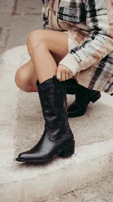 Bota de cuero negro estilo texana con caña media. Presenta un aplique desmontable en la zona del tobillo decorado con tachas metálicas y flecos largos. Posee taco bajo y punta levemente afinada.