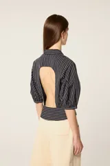 Camisa corta de popelina a rayas con mangas abullonadas, cuello solapa y cierre frontal con botón decorativo. Presenta un diseño con efecto plisado en el pecho y corte ajustado.