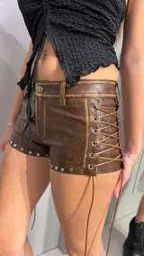 Short de cuero color marrón, tiro bajo, con tachas metálicas en el borde inferior y cordones entrelazados en los laterales.