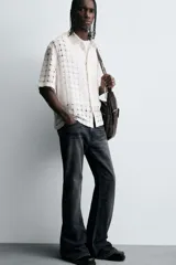 Camisa negra de corte relajado con tejido semitransparente de estructura abierta y patrón a cuadros. Presenta cuello solapa, manga corta, bolsillo plastrón en el pecho y cierre frontal de botones.