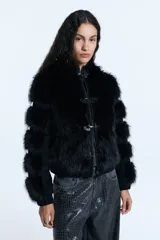 Campera de pelo ecológico negro con recortes en denim negro. Presenta cierre metálico frontal, alamares metálicos decorativos, capucha y bolsillos laterales. Las mangas tienen un diseño con recortes que alternan pelo y denim, y cuenta con elástico en la cintura.